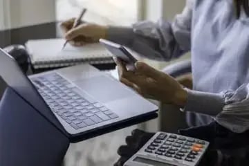 man with phone at laptop writing on a notepad